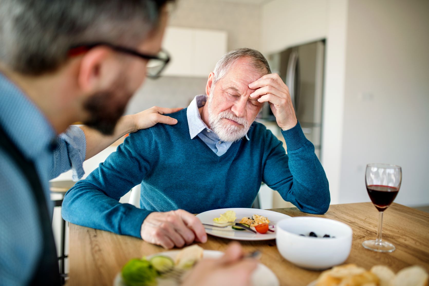 Mature man sitting at a table frustrated with son whit his hand on his shoulder